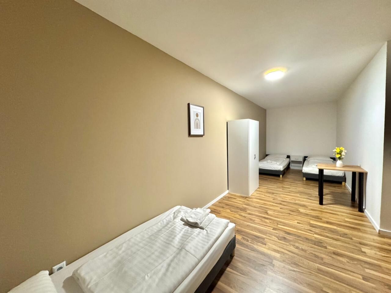 A compact room with a single bed, a small table with a vase, and a door leading to another room.