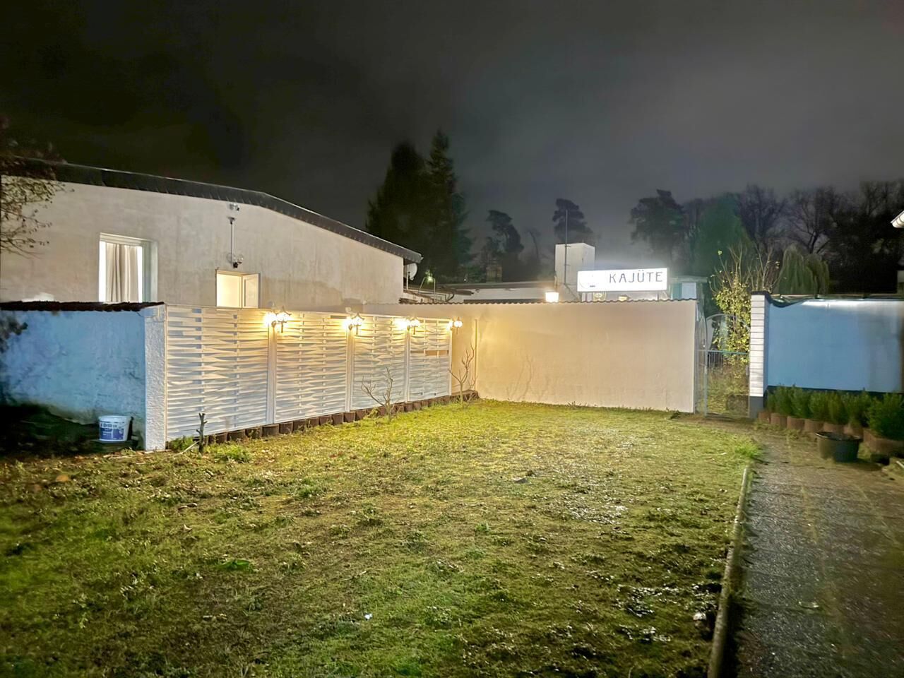 Een goed verlicht buitengebied met een wit gebouw, een overdekte zithoek en een bordje met de tekst 'KAJUTE'.