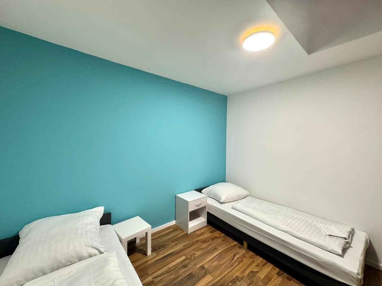 A small, single-room accommodation with two beds, a nightstand, and a ceiling light.