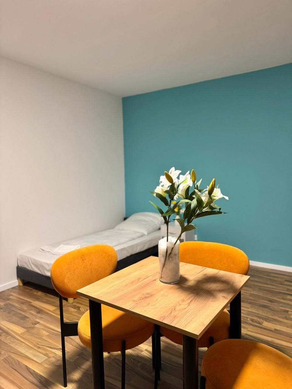 A small room with a wooden table, two orange chairs, a white vase with flowers, and a single bed against a teal wall.