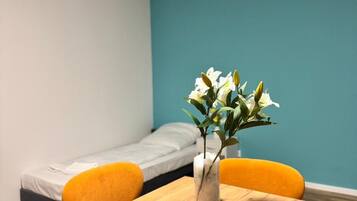 A small room with a wooden table, two orange chairs, a white vase with flowers, and a single bed against a teal wall.