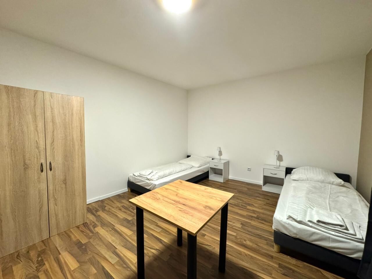 Een kleine, schone kamer met twee enkele bedden, een houten garderobe, een klein tafeltje en een nachtkastje met een lamp.