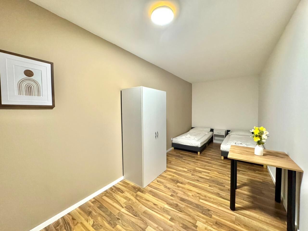 A compact room with a bed, a small table with a vase, a wardrobe, and a framed picture on the wall.