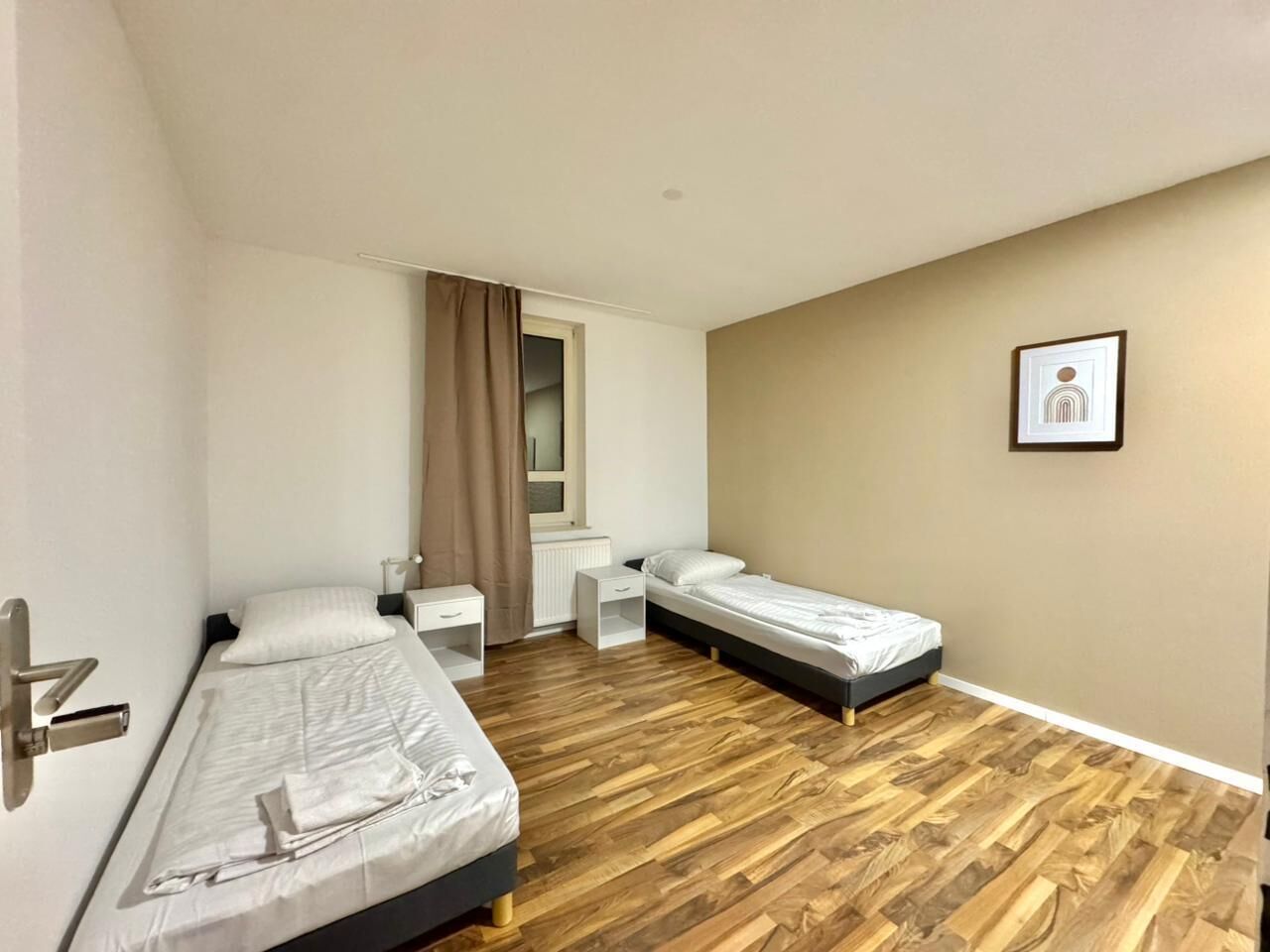 Una habitación pequeña y limpia con dos camas individuales, una mesita, una silla y una ventana con cortinas.