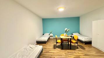 Une petite pièce bien éclairée, avec trois lits simples, un plancher en bois, une table avec des chaises et un mur d’accent bleu.