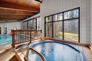 An indoor swimming pool with a wooden deck, large windows, and a view of the outdoors.