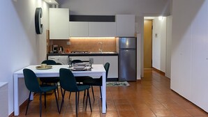 A compact kitchen with white cabinets, a stainless steel refrigerator, and a dining area with a white table and green chairs. - Veronetta 52 Garden - Verona Journeys (Verona)
