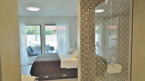 A bedroom with a bed, a bedside table, a shower area, and a view of the outside.