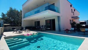 A modern two-story house with a swimming pool, outdoor seating, and a clear blue sky.