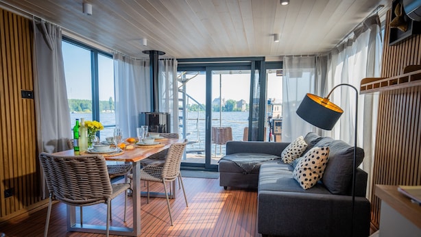 A modern interior with a dining area, sofa, and large windows overlooking a body of water. - Floatinghouse Aquila WOMA D13 – Modern Houseboat for 6 Guests with Rooftop Terrace (Fürstenberg)