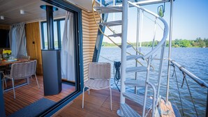 A deck with a table and chairs, a staircase, and a view of the water. - Floatinghouse Aquila WOMA D13 – Modern Houseboat for 6 Guests with Rooftop Terrace (Fürstenberg)
