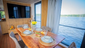 A dining table set with plates, glasses, and a vase of yellow flowers, overlooking a body of water. - Floatinghouse Aquila WOMA D13 – Modern Houseboat for 6 Guests with Rooftop Terrace (Fürstenberg)