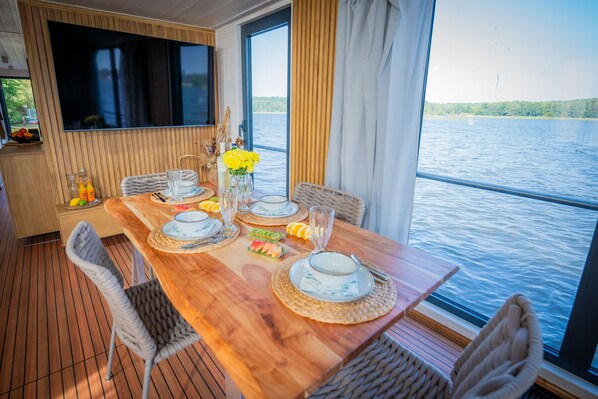 Une table à manger dressée avec des assiettes, des verres et un vase rempli de fleurs jaunes, donnant sur une étendue d’eau.
