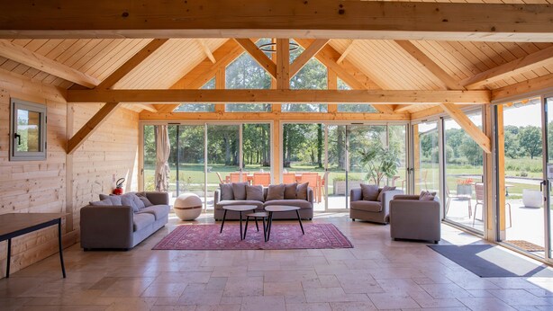 Une pièce spacieuse avec un plafond haut, des poutres en bois, de grandes fenêtres et un coin salon comprenant des canapés et une table basse.