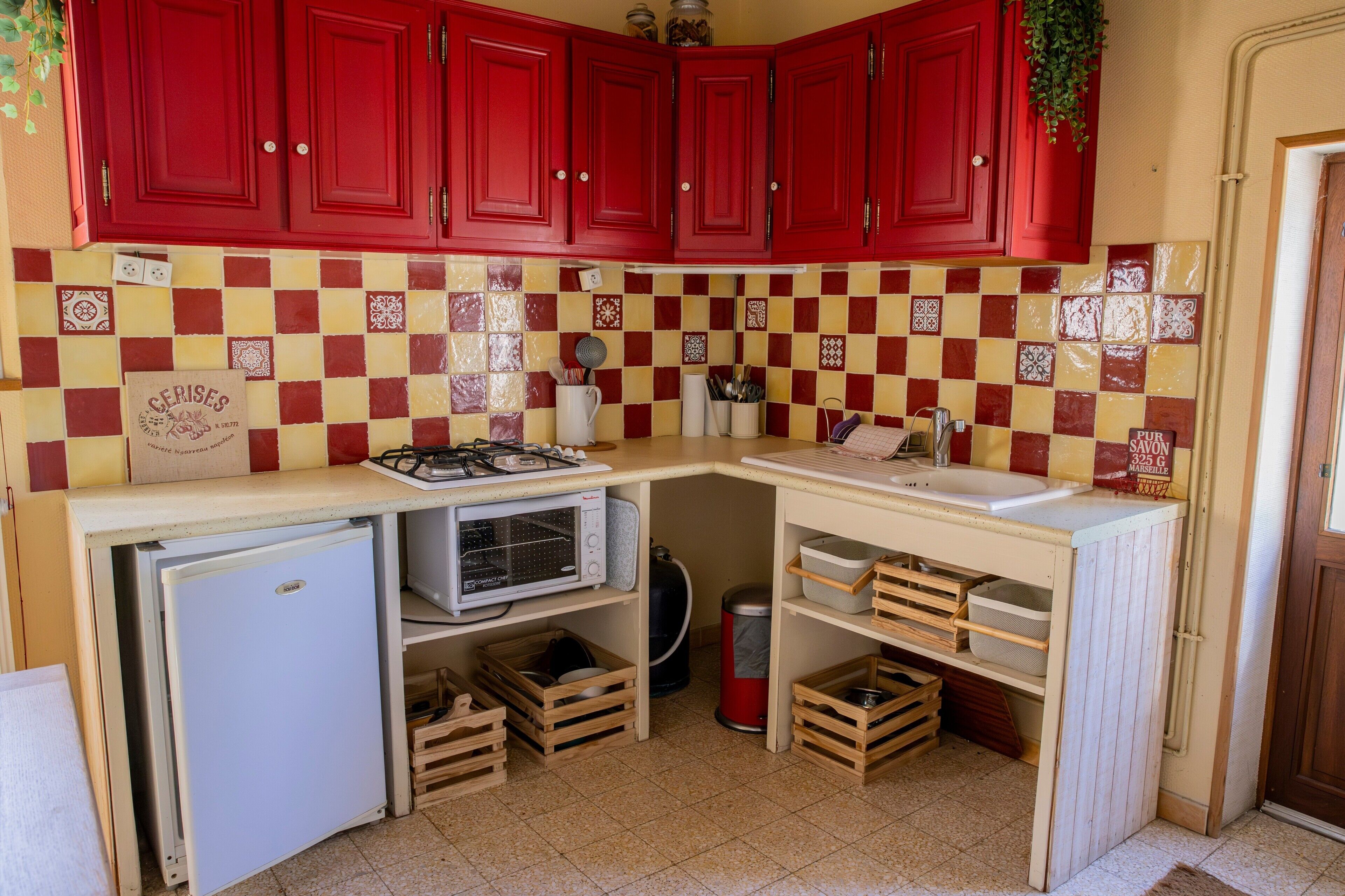 Een kleine keuken met rode kastjes, een magnetron, een spoelbak en opbergkisten.