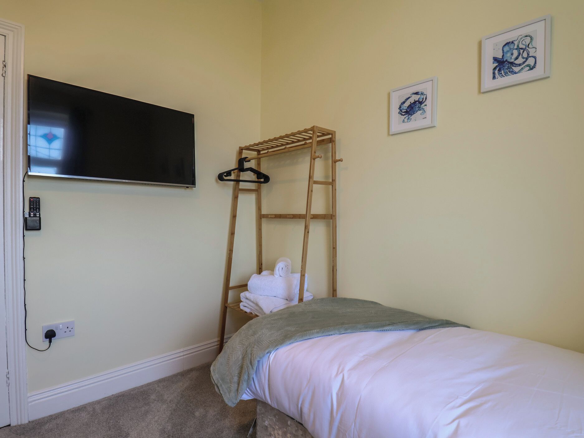 Un dormitorio con cama, televisor montado en la pared, escalera de madera, perchero y dos cuadros enmarcados.