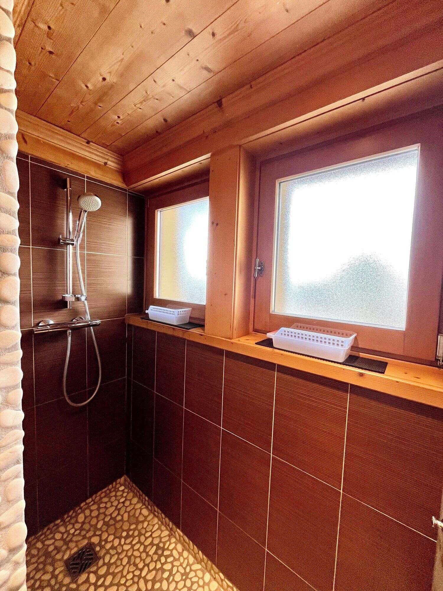 Un baño rústico con cabaña, con ducha, dos ventanas y piso de baldosas.