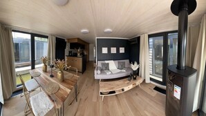 A modern living space with a dining area, a sofa, a fireplace, and a kitchen in the background. - Floatinghouse Enjoy WOMA D13 – Modernes Hausboot mit Dachterrasse am Röblinsee (Fürstenberg)