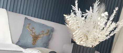 A modern living room with a grey sofa, a decorative sculpture, a tall vase with white foliage, and a framed artwork on the wall.