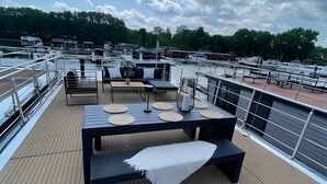 A deck with outdoor seating, a table, and a view of boats and greenery. - Floatinghouse Enjoy WOMA D13 – Modernes Hausboot mit Dachterrasse am Röblinsee (Fürstenberg)