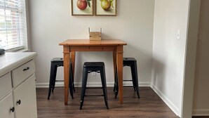 Ein kleiner Essbereich mit einem Holztisch und zwei schwarzen Hocker, einem weißen Schrank und zwei gerahmten Bildern von Äpfeln an der Wand.