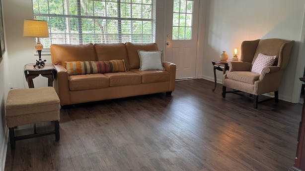 Ein Wohnzimmer mit einem braunen Ledersofa, einem passenden Sessel und einem kleinen Hocker. Es gibt ein Fenster mit Jalousien, eine Tür und eine Lampe auf einem Beistelltisch.