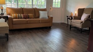 Ein Wohnzimmer mit braunem Ledersofa, Holzboden, Deckenventilator und einem Fenster mit Jalousien.
