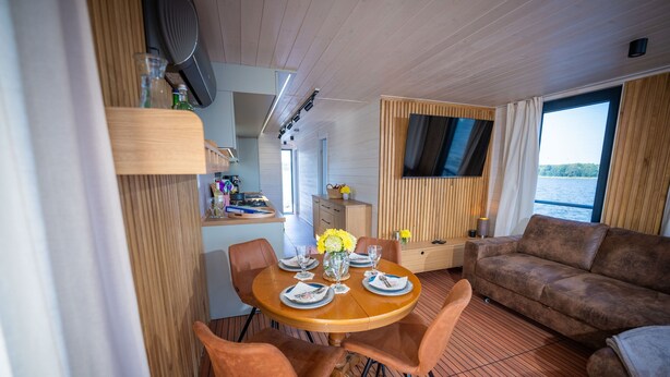 A dining area with a round table, chairs, and a television on the wall. - Floatinghouse Frigga WOMA D13 – Modernes Hausboot mit Dachterrasse am Röblinsee (Fürstenberg)