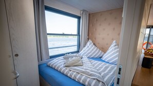 A bedroom with a window, a bed with striped bedding, and a view of the water outside. - Floatinghouse Frigga WOMA D13 – Modernes Hausboot mit Dachterrasse am Röblinsee (Fürstenberg)