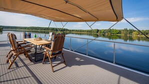A deck with a table and chairs, a canopy, and a lake view. - Floatinghouse Frigga WOMA D13 – Modernes Hausboot mit Dachterrasse am Röblinsee (Fürstenberg)