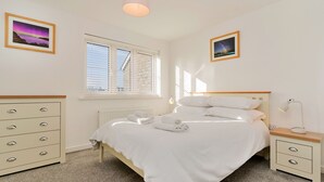 Een slaapkamer met een bed, nachtkastjes, een dressoir, een raam met jaloezieën en twee ingelijste foto's aan de muur.
