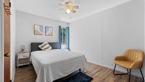 Ein Schlafzimmer mit Bett, Nachttisch, Stuhl, Deckenventilator und einem Fenster mit Vorhängen.