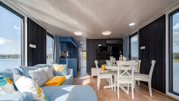 A modern interior with a dining area, a sofa with cushions, and large windows offering a view of the water.