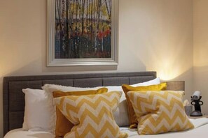Una cama con cabecera gris, almohadas blancas y amarillas con estampado en zigzag, y un cuadro abstracto enmarcado encima.