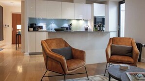 A modern kitchen with white cabinets, a central island, and two leather armchairs. - Sunny Westminster 1-Bed with Terrace & AC – Walk to London’s Icons (London)