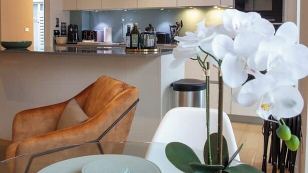 A modern kitchen with a central island, a dining area with a glass table, and a white orchid plant. - Sunny Westminster 1-Bed with Terrace & AC – Walk to London’s Icons (London)
