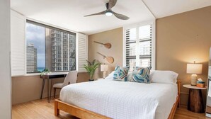 A bedroom with a bed, a desk, a chair, a ceiling fan, and a view of buildings through the window. - Chic Suite W/ Direct Ocean Views & Free Parking (Honolulu)