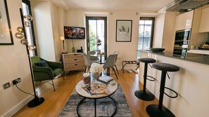 A modern living room with a green sofa, a coffee table with magazines, and a kitchen area with a counter and stools. - Elegant Westminster Retreat: Stylish 2-Bed, 2-Bath with Air-Con (London)