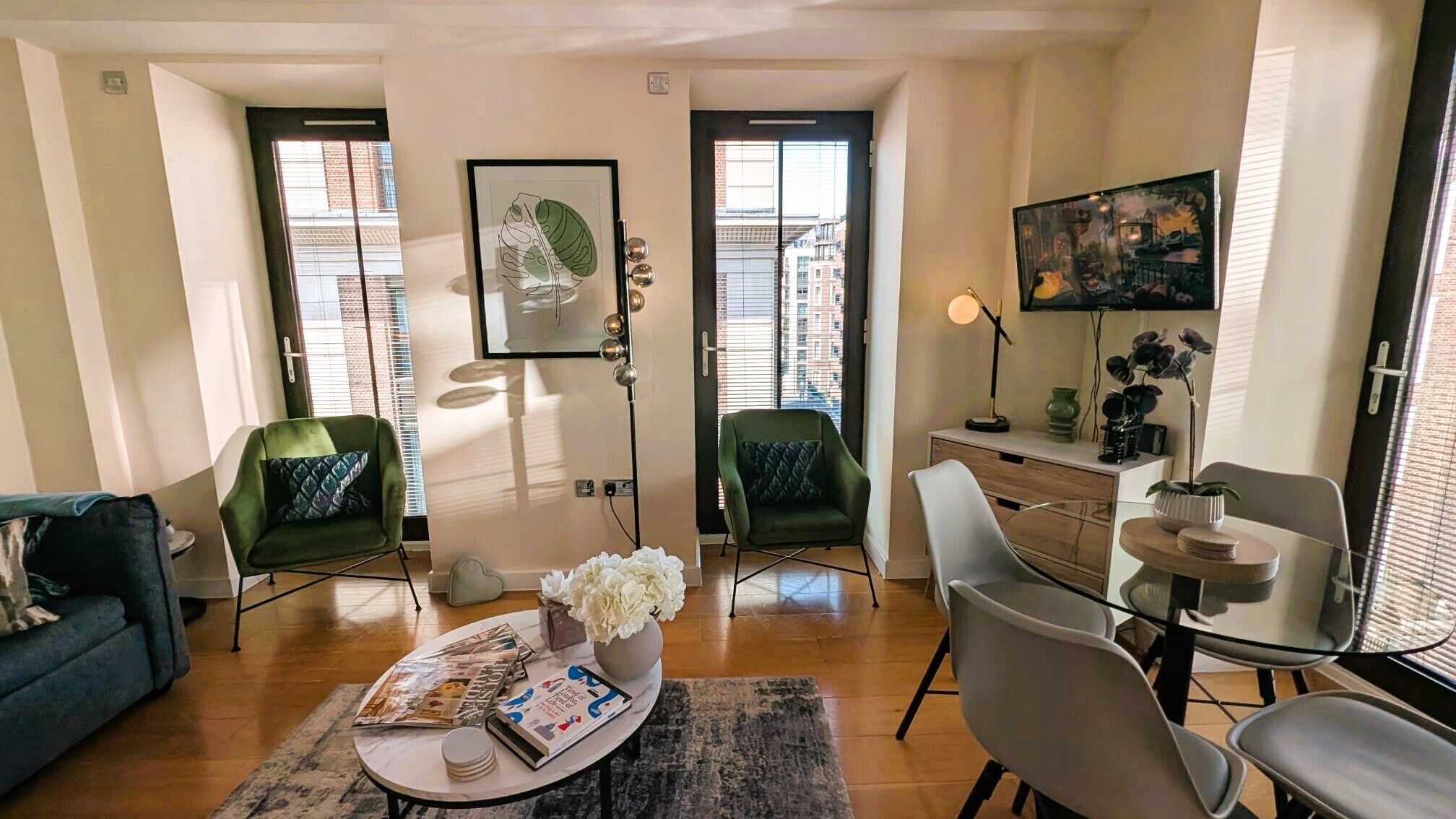 Un salon comprenant un canapé, deux fauteuils, une table basse avec des magazines, un vase de fleurs et un bureau avec une lampe et un tableau.