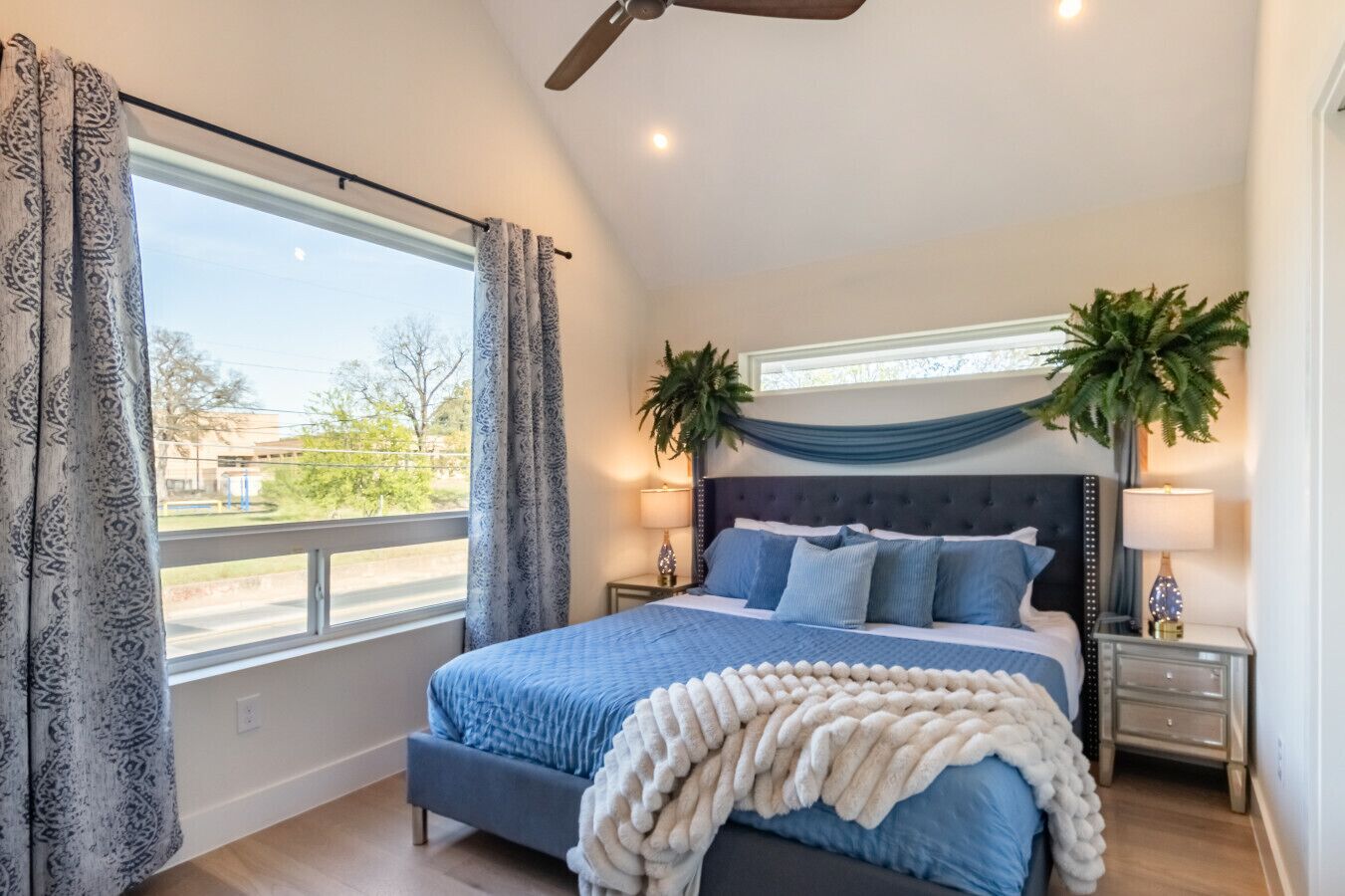 A bedroom with a large bed, two bedside tables, a ceiling fan, and a view of the outdoors.