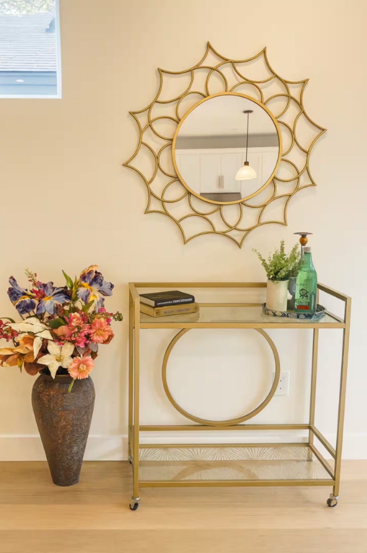 A room with a decorative wall mirror, a vase of flowers, and a metal console table with a book and a bottle.