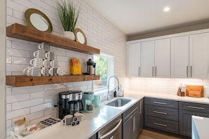 A modern kitchen with white subway tiles, wooden shelves, and grey cabinets. - Luxe 6BR Oasis | Pool•Hot Tub•Games | Near DTown (Austin)