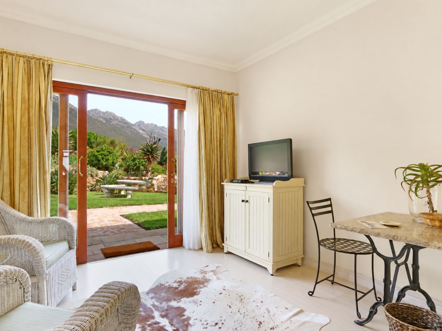 Una habitación con televisión en un mueble, una mesa de comedor con sillas, un sofá y vistas a un jardín y montañas a través de un ventanal corredero.