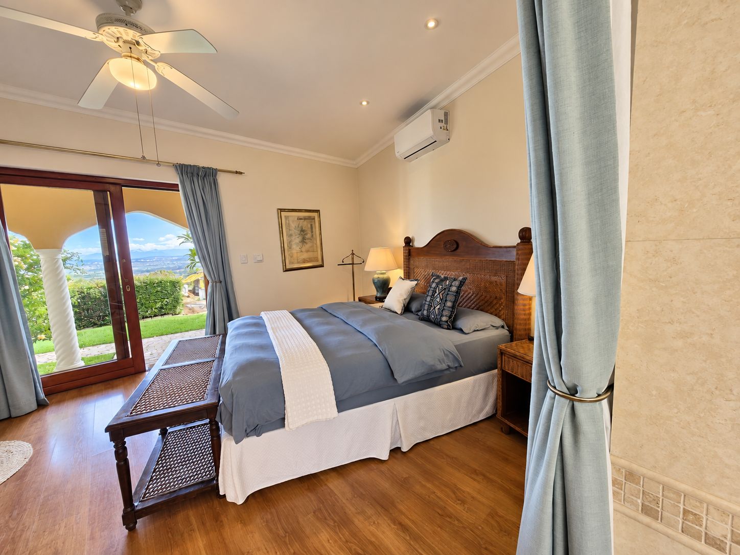 A bedroom with a bed, bedside tables, a ceiling fan, an air conditioning unit, and a view through a glass door.