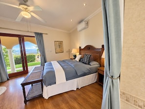 Une chambre à coucher comprenant un lit, des tables de chevet, un ventilateur de plafond, un appareil de climatisation et une vue sur l’extérieur, visible à travers une porte vitrée.