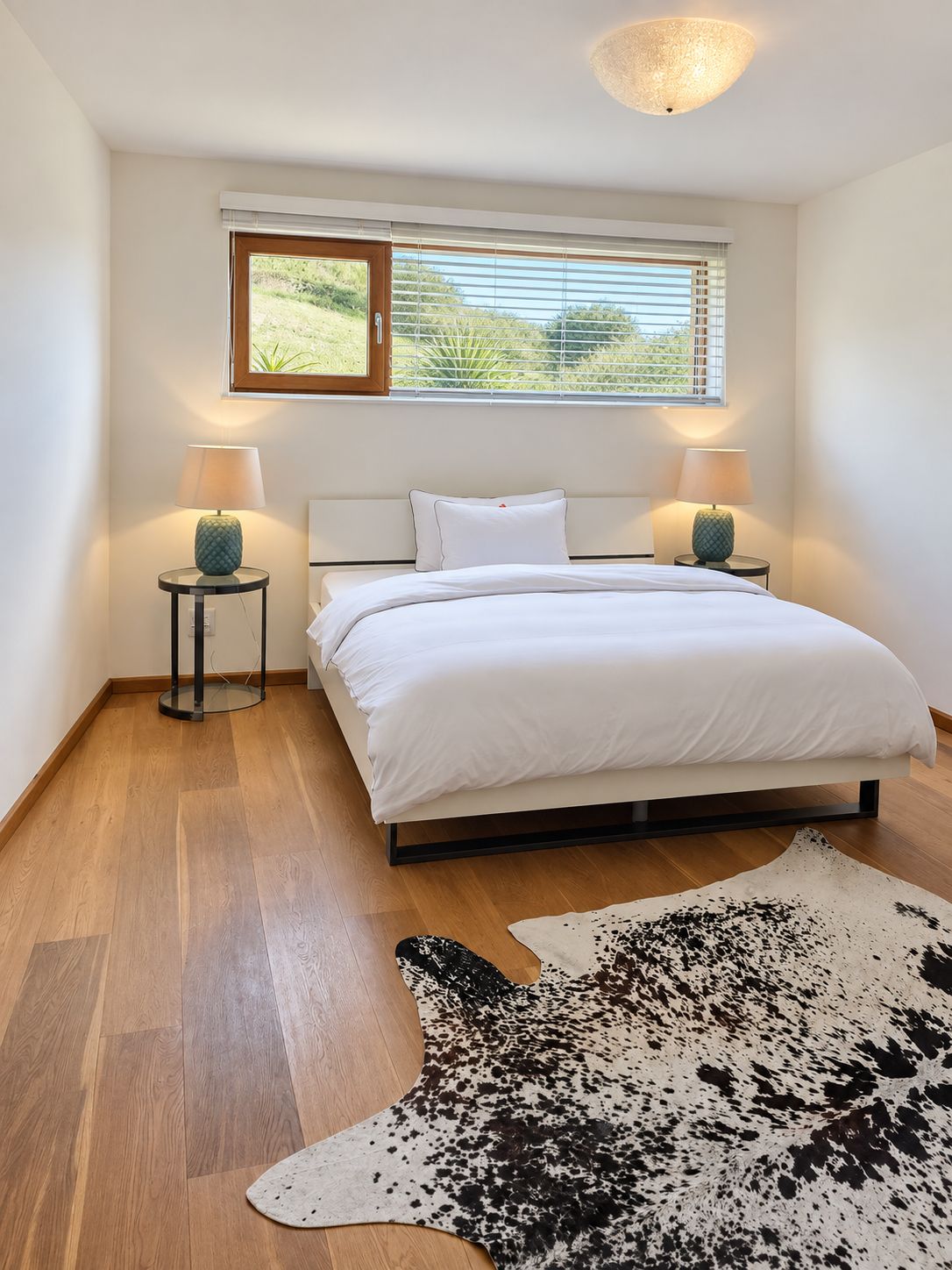A bedroom with a bed, two bedside lamps, a window with blinds, and a cowhide rug on the floor.