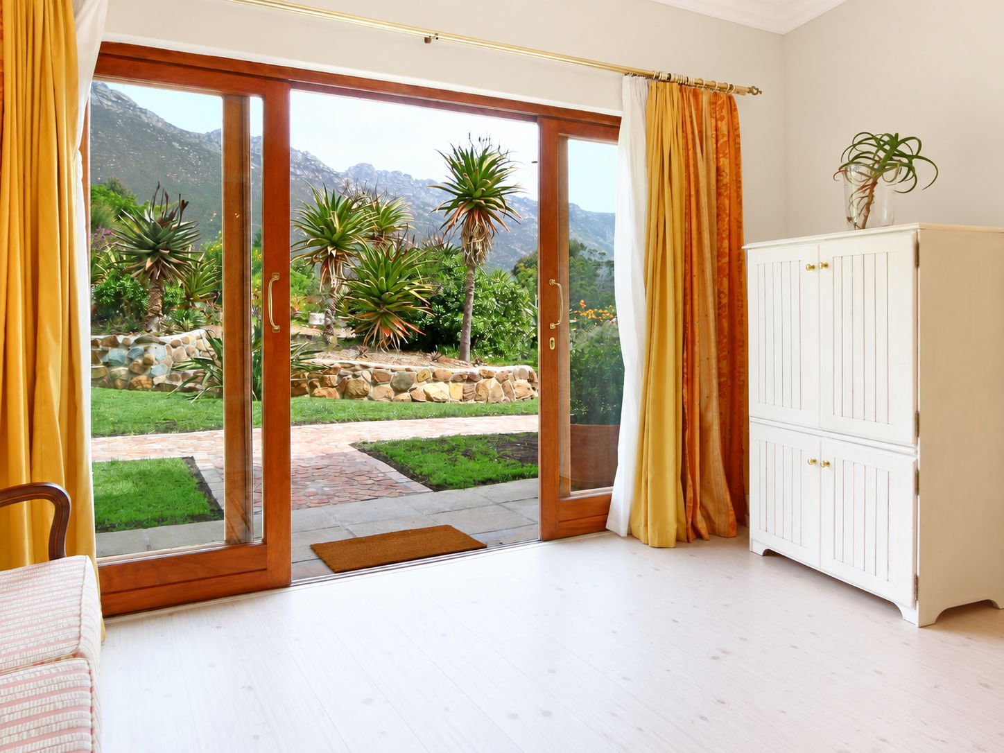 A room with a sliding glass door leading to a garden, a white cabinet, and a plant on a table.