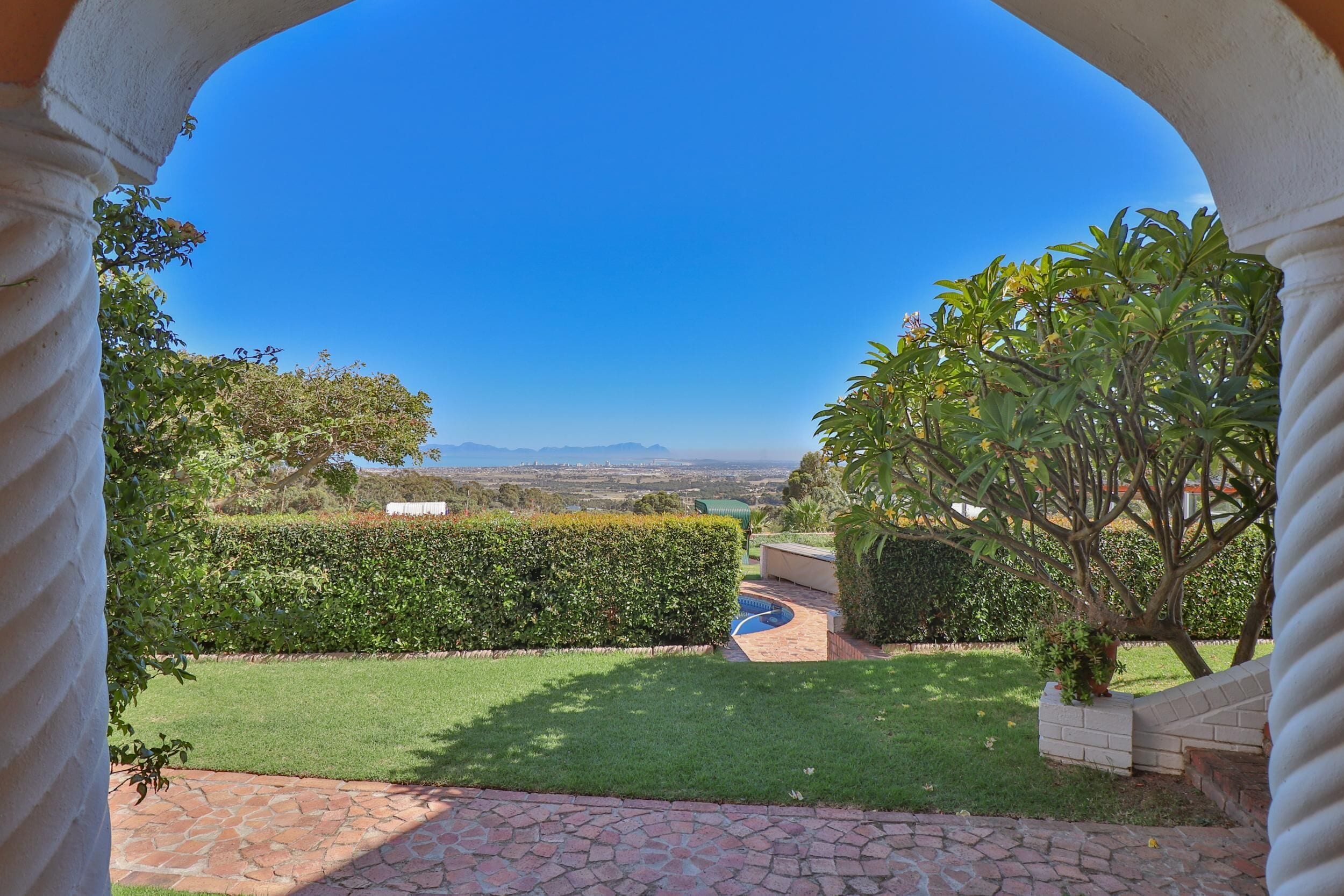 A view through an archway of a well-maintained garden with a clear sky, a hedge, and a brick pathway leading to a pool.