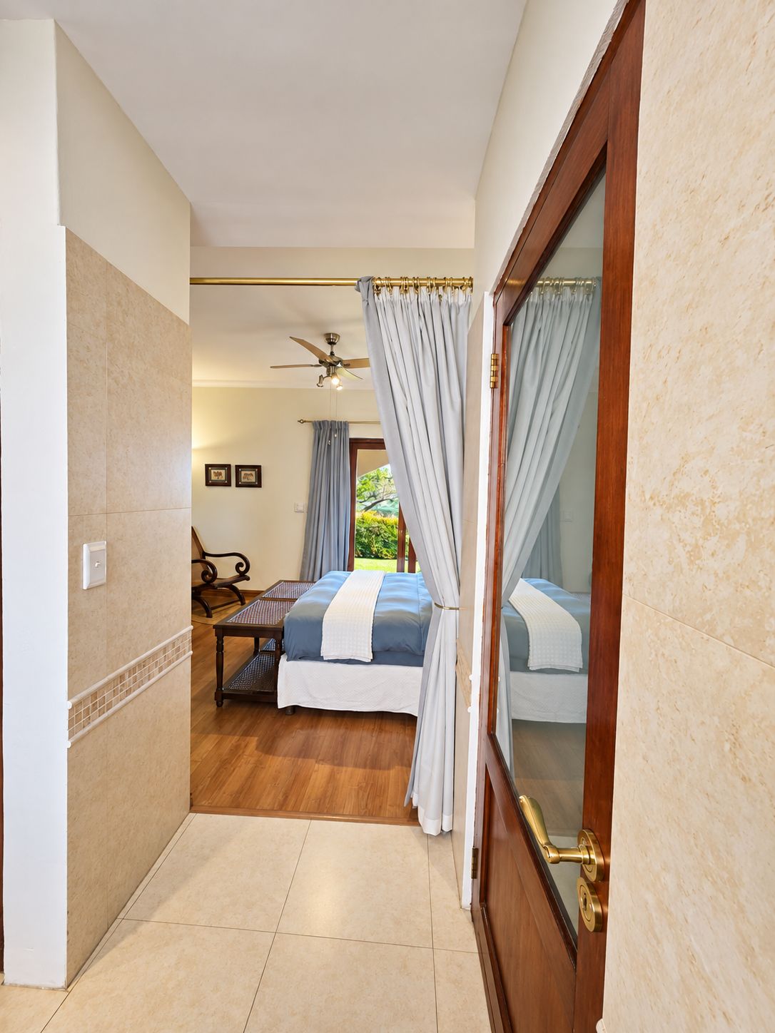 Una habitación de hotel con una cama, un ventilador de techo, una ventana con cortinas y una puerta de madera.