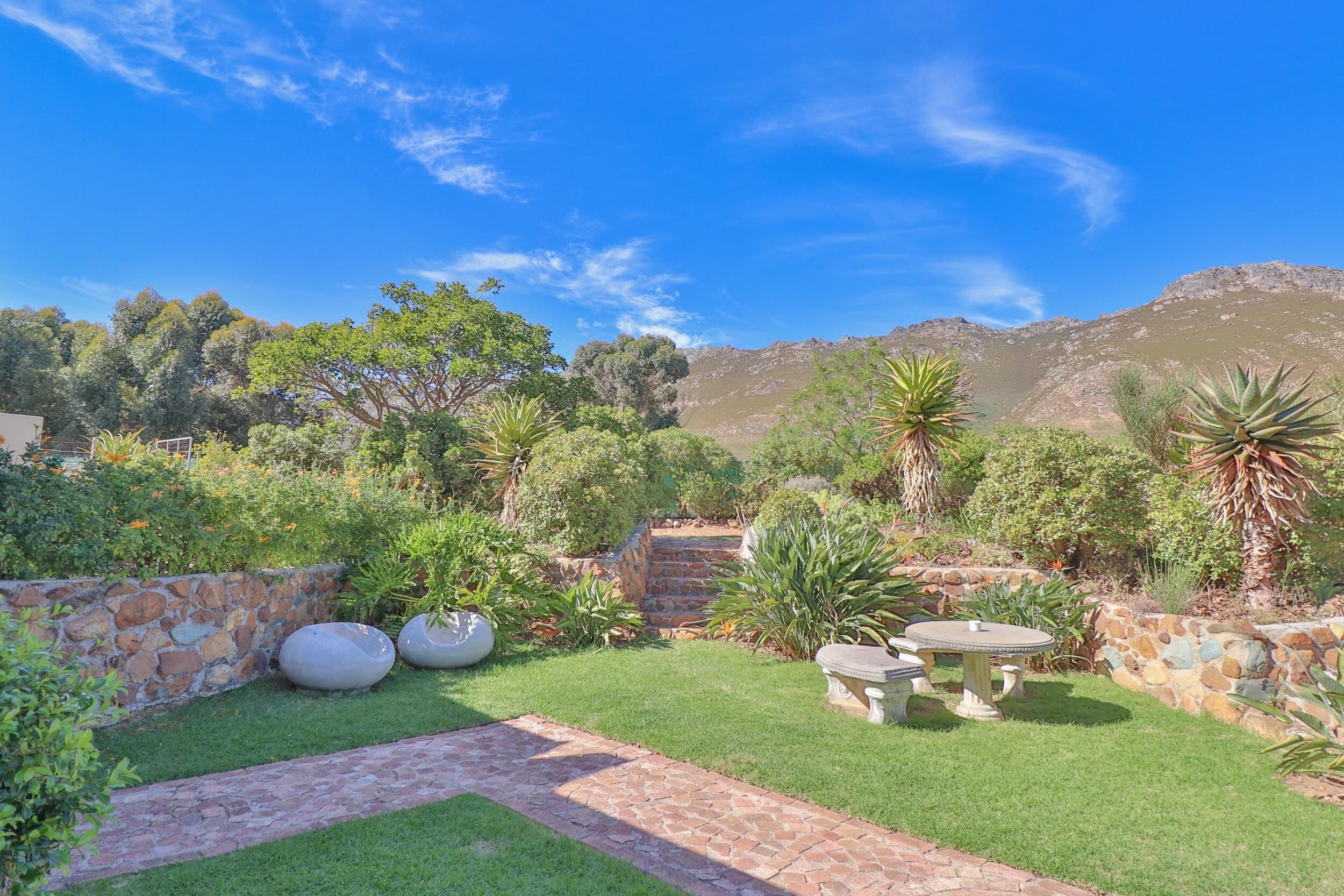 A landscaped garden with a stone wall, a paved pathway, and a seating area with a table and chairs.